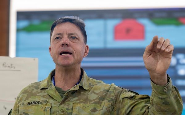 An Australian military officer named Hardy is delivering a briefing or presentation in what appears to be a command or operations room. He is gesturing with his right hand while holding a marker in his left, standing in front of a whiteboard and a large display screen showing a map or operational graphic. The setting suggests a military planning or training exercise environment.