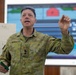 An Australian military officer named Hardy is delivering a briefing or presentation in what appears to be a command or operations room. He is gesturing with his right hand while holding a marker in his left, standing in front of a whiteboard and a large display screen showing a map or operational graphic. The setting suggests a military planning or training exercise environment.
