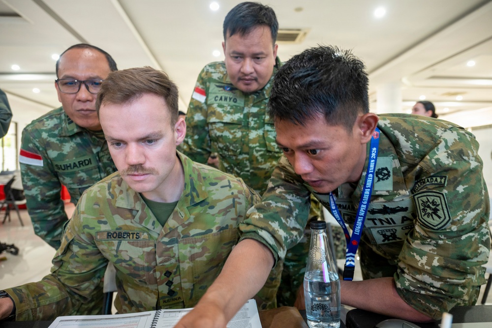 Military personnel from multiple nations are gathered around a table reviewing documents or a laptop in what appears to be an indoor briefing or planning session. An Australian soldier identified as 'Roberts' is seated at the center, surrounded by Indonesian Army (TNI AD) soldiers including individuals named 'Suhardi' and 'Canyo'. The setting and lanyards suggest this is part of a multinational military exercise, specifically 'Latgabma Super Garuda Shield Tahun 2026'.