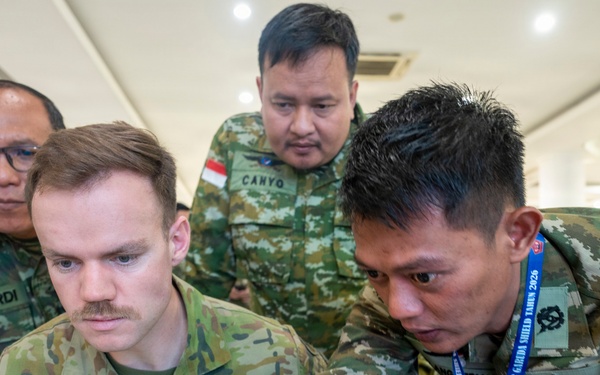 Military personnel from multiple nations are gathered around a table reviewing documents or a laptop in what appears to be an indoor briefing or planning session. An Australian soldier identified as 'Roberts' is seated at the center, surrounded by Indonesian Army (TNI AD) soldiers including individuals named 'Suhardi' and 'Canyo'. The setting and lanyards suggest this is part of a multinational military exercise, specifically 'Latgabma Super Garuda Shield Tahun 2026'.