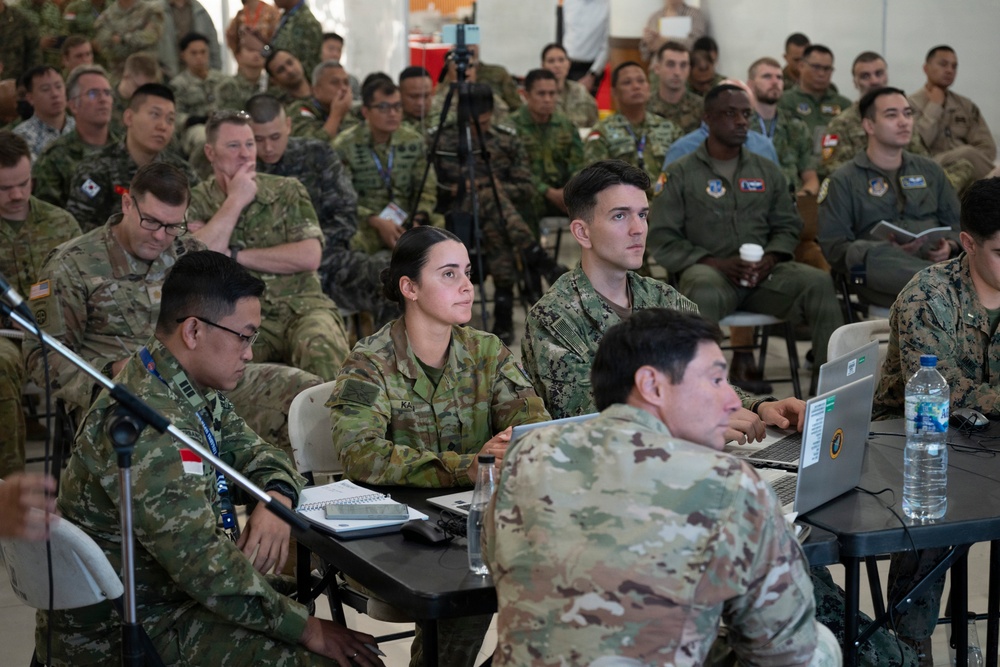 A large multinational military briefing or conference is taking place in what appears to be a tent or temporary facility. Personnel from multiple nations are seated in rows, wearing various military uniforms including US Army, US Navy, US Air Force, Australian Army, and other allied nation uniforms. Several attendees at front tables are working on laptops while the group attends a presentation.