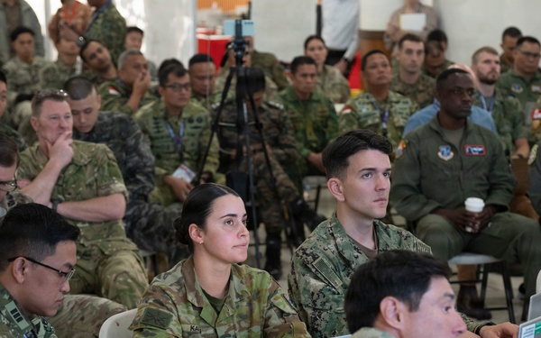 A large multinational military briefing or conference is taking place in what appears to be a tent or temporary facility. Personnel from multiple nations are seated in rows, wearing various military uniforms including US Army, US Navy, US Air Force, Australian Army, and other allied nation uniforms. Several attendees at front tables are working on laptops while the group attends a presentation.