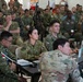 A large multinational military briefing or conference is taking place in what appears to be a tent or temporary facility. Personnel from multiple nations are seated in rows, wearing various military uniforms including US Army, US Navy, US Air Force, Australian Army, and other allied nation uniforms. Several attendees at front tables are working on laptops while the group attends a presentation.