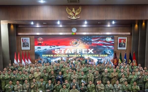 A large group of military personnel from multiple nations pose for a group photograph in a formal conference room. The backdrop displays a banner reading 'STAFFEX - Super Garuda Shield Exercise 2026,' indicating this is a joint military staff exercise. Indonesian and American flags are visible alongside the Indonesian national emblem (Garuda Pancasila), suggesting a bilateral or multinational military cooperation event.