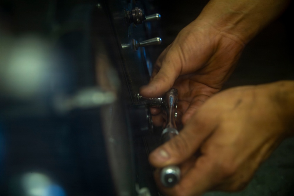 A close-up photograph of a person's hands working on a mechanical component, likely a drum or similar instrument hardware. The hands appear to be adjusting or tightening metal hardware using a small tool. The image is taken in low-light conditions with shallow depth of field.