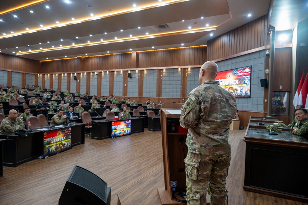 A military briefing or presentation is taking place in a formal auditorium-style conference room. A soldier in MultiCam/OCP camouflage uniform stands at a podium addressing an audience of military personnel seated in rows. Signage reading 'STAFFEX' is visible on screens and banners, indicating this is a staff exercise event, likely a joint U.S.-Indonesian military exercise based on the visible flags.