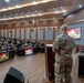 A military briefing or presentation is taking place in a formal auditorium-style conference room. A soldier in MultiCam/OCP camouflage uniform stands at a podium addressing an audience of military personnel seated in rows. Signage reading 'STAFFEX' is visible on screens and banners, indicating this is a staff exercise event, likely a joint U.S.-Indonesian military exercise based on the visible flags.
