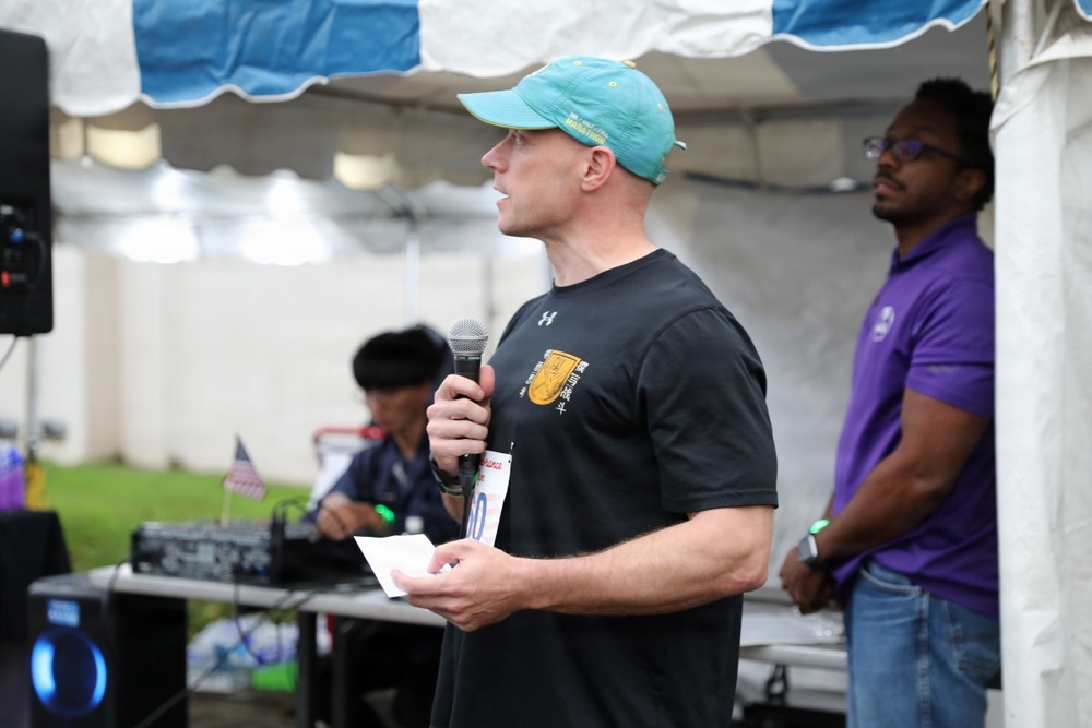 A man wearing a teal baseball cap and black Under Armour t-shirt is speaking into a microphone at an outdoor event held under a tent. He is holding a piece of paper, suggesting he may be reading from notes or an announcement. In the background, a DJ setup is visible along with another individual in a purple shirt.