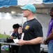 A man wearing a teal baseball cap and black Under Armour t-shirt is speaking into a microphone at an outdoor event held under a tent. He is holding a piece of paper, suggesting he may be reading from notes or an announcement. In the background, a DJ setup is visible along with another individual in a purple shirt.