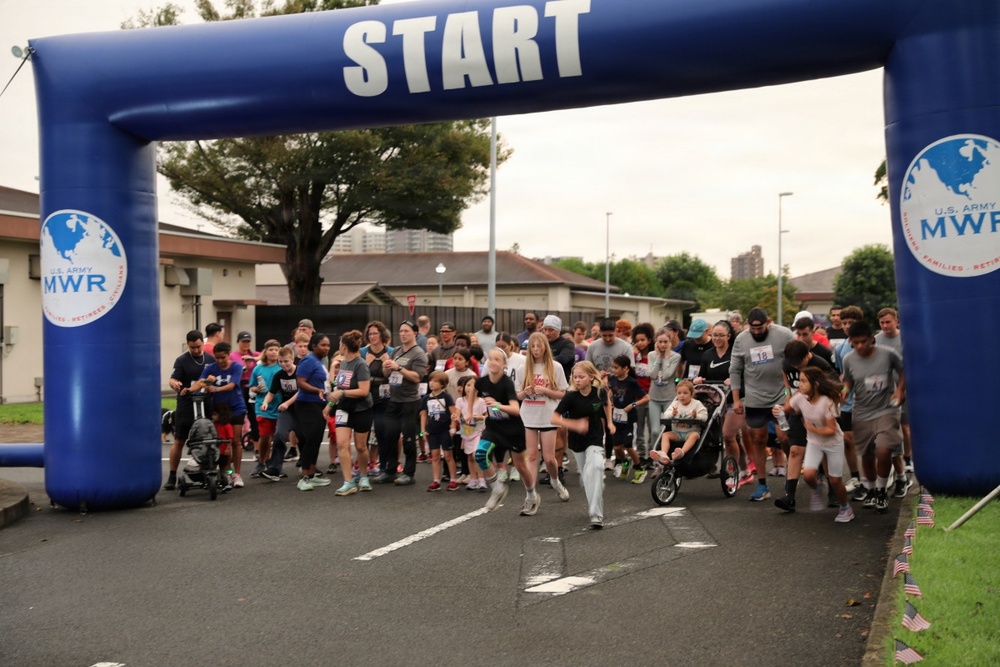 A large group of participants, including adults and children, are crossing the start line of a fun run or 5K race organized by U.S. Army MWR (Morale, Welfare, and Recreation). The event appears to be held on a military installation, with participants of all ages including families with strollers. The overcast sky and casual attire suggest a community fitness event rather than a competitive race.