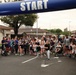A large group of participants, including adults and children, are crossing the start line of a fun run or 5K race organized by U.S. Army MWR (Morale, Welfare, and Recreation). The event appears to be held on a military installation, with participants of all ages including families with strollers. The overcast sky and casual attire suggest a community fitness event rather than a competitive race.