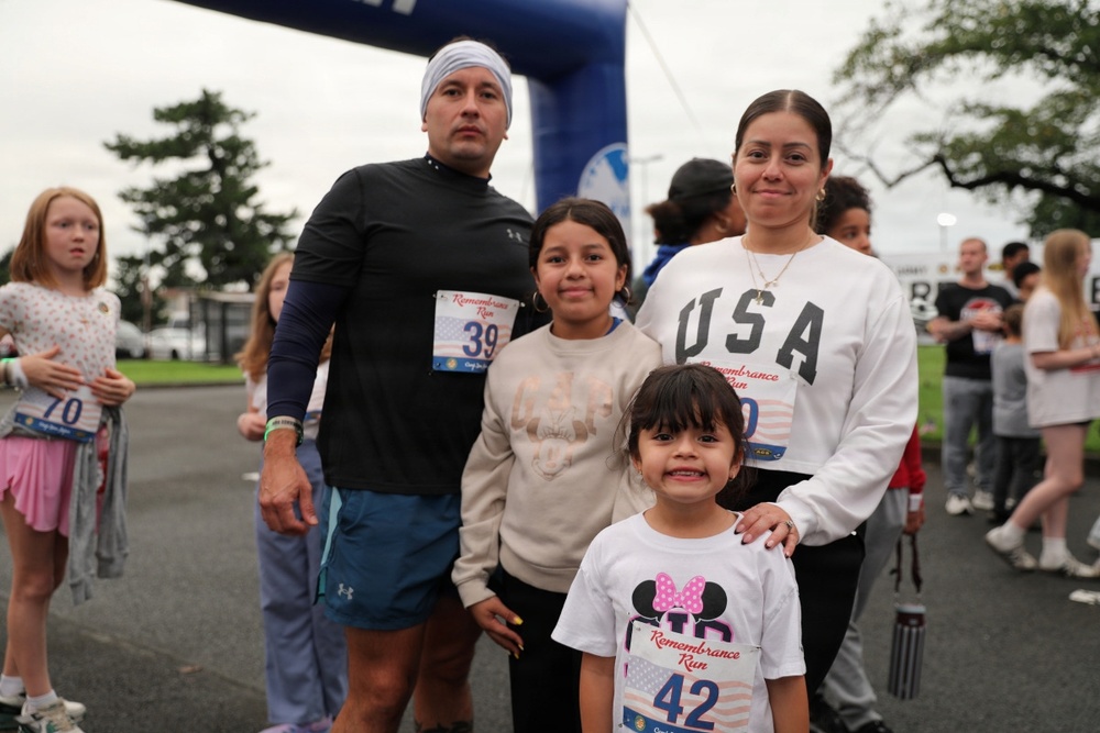 A family of four poses together at a 'Remembrance Run' event, standing near the starting/finishing arch. The group consists of two adults and two children, all wearing race bibs. The event appears to be a community running event held outdoors on an overcast day.
