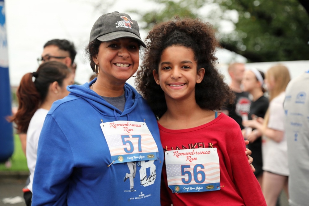 Two smiling individuals — an adult woman and a young girl — pose together at an outdoor running event called the 'Remembrance Run.' The woman wears bib number 57 and the girl wears bib number 56, both featuring an American flag design. Several other participants are visible in the background, suggesting a community race or charity run.