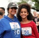 Two smiling individuals — an adult woman and a young girl — pose together at an outdoor running event called the 'Remembrance Run.' The woman wears bib number 57 and the girl wears bib number 56, both featuring an American flag design. Several other participants are visible in the background, suggesting a community race or charity run.