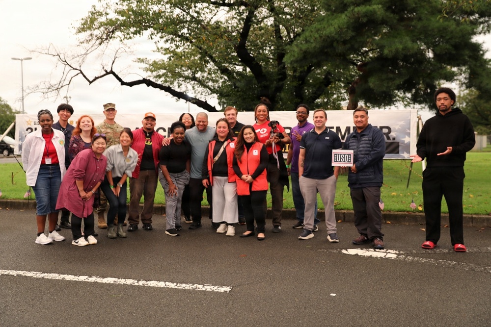 A group of approximately 20 people pose together outdoors in a parking lot area for a group photo at what appears to be a USO event. A large banner is visible behind them with partial text reading 'REMEMBER,' and one person is holding a USO sign. The group is diverse and casually dressed, with one individual in military camouflage uniform.