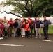 A group of approximately 20 people pose together outdoors in a parking lot area for a group photo at what appears to be a USO event. A large banner is visible behind them with partial text reading 'REMEMBER,' and one person is holding a USO sign. The group is diverse and casually dressed, with one individual in military camouflage uniform.