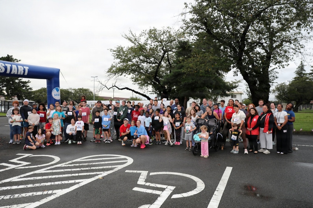 A large group of participants, including adults and children of various ages, pose together at the start line of a 5K run/race event. The group is gathered under a blue inflatable 'START' arch, with many participants wearing race bib numbers. The event appears to be a community or military-affiliated fun run, likely organized by MWR (Morale, Welfare, and Recreation).