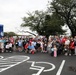 A large group of participants, including adults and children of various ages, pose together at the start line of a 5K run/race event. The group is gathered under a blue inflatable 'START' arch, with many participants wearing race bib numbers. The event appears to be a community or military-affiliated fun run, likely organized by MWR (Morale, Welfare, and Recreation).