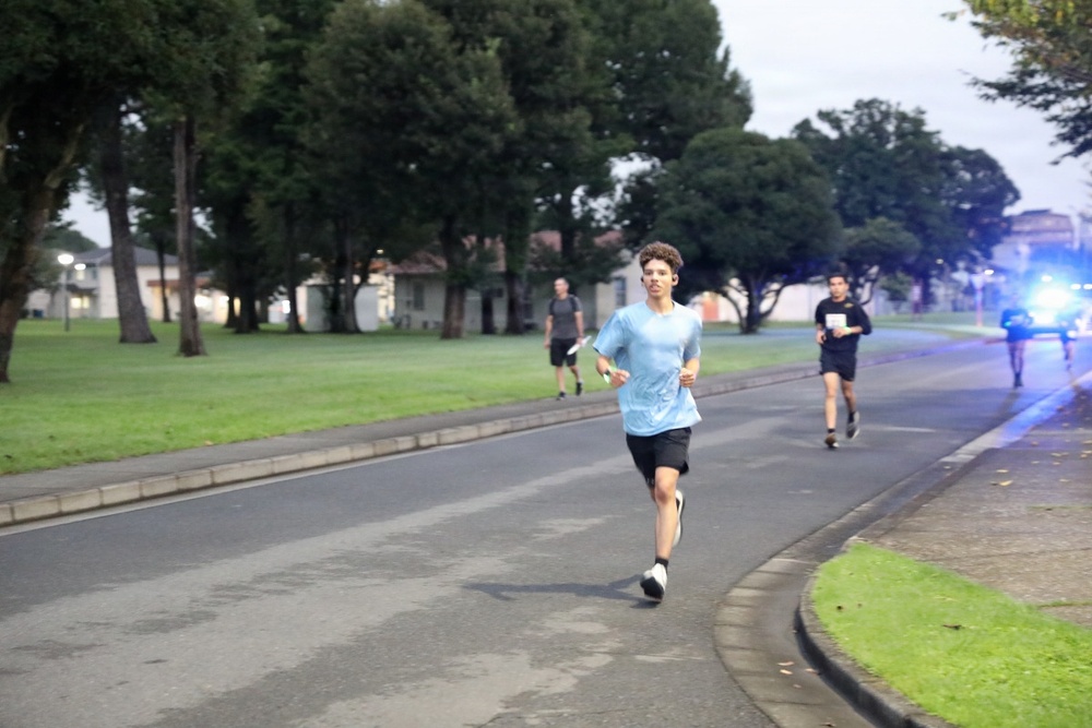 A group of runners are jogging along a road in what appears to be a park or institutional campus during early morning or evening low-light conditions. The lead runner is a young male wearing a light blue t-shirt and black shorts. Blue emergency or escort vehicle lights are visible in the background, suggesting this may be an organized run or fitness event.