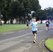 A group of runners are jogging along a road in what appears to be a park or institutional campus during early morning or evening low-light conditions. The lead runner is a young male wearing a light blue t-shirt and black shorts. Blue emergency or escort vehicle lights are visible in the background, suggesting this may be an organized run or fitness event.