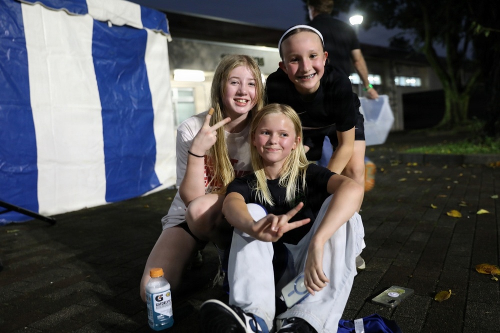 Three young girls pose together outdoors at night, smiling and making peace signs for the camera. They appear to be at an outdoor event, evidenced by a blue and white tent/canopy visible in the background. The setting appears to be a paved area near a building, likely a school or community facility.