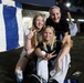 Three young girls pose together outdoors at night, smiling and making peace signs for the camera. They appear to be at an outdoor event, evidenced by a blue and white tent/canopy visible in the background. The setting appears to be a paved area near a building, likely a school or community facility.