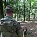 A military or law enforcement handler named 'BUSIC' wearing OCP/MultiCam camouflage uniform and plate carrier vest stands in a wooded area facing a Belgian Malinois military working dog. The dog is seated attentively in front of the handler, suggesting a training or patrol exercise. The setting appears to be a forested training area during daylight hours.