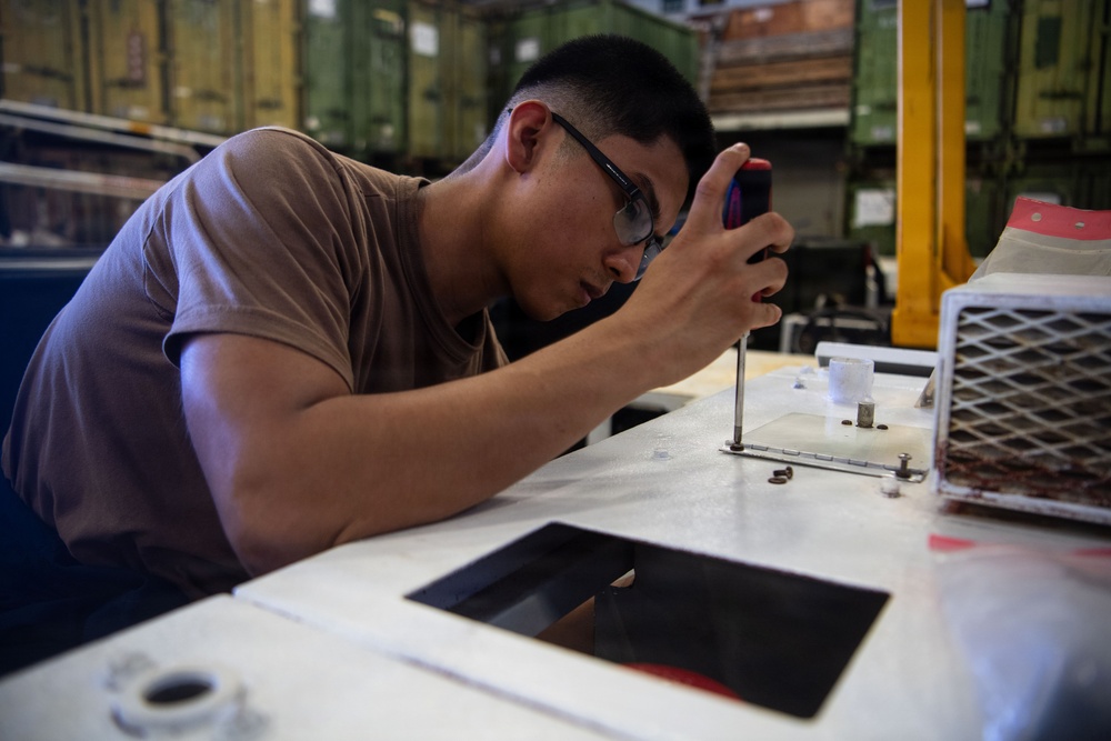 A young man wearing safety glasses and a tan/brown t-shirt is using a screwdriver to work on a mechanical or electronic component mounted on a white surface. He is leaning in closely for precision work in what appears to be an industrial or military maintenance environment. Small screws and hardware are visible on the work surface nearby.