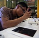 A young man wearing safety glasses and a tan/brown t-shirt is using a screwdriver to work on a mechanical or electronic component mounted on a white surface. He is leaning in closely for precision work in what appears to be an industrial or military maintenance environment. Small screws and hardware are visible on the work surface nearby.