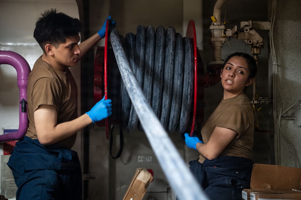 Two military personnel in matching brown t-shirts and dark work pants are handling a large fire hose reel with a red frame in what appears to be a shipboard compartment. Both individuals are wearing blue nitrile gloves and appear to be either deploying or recoiling the hose. The industrial setting with visible piping and valves in the background is consistent with a naval vessel interior.