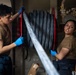Two military personnel in matching brown t-shirts and dark work pants are handling a large fire hose reel with a red frame in what appears to be a shipboard compartment. Both individuals are wearing blue nitrile gloves and appear to be either deploying or recoiling the hose. The industrial setting with visible piping and valves in the background is consistent with a naval vessel interior.