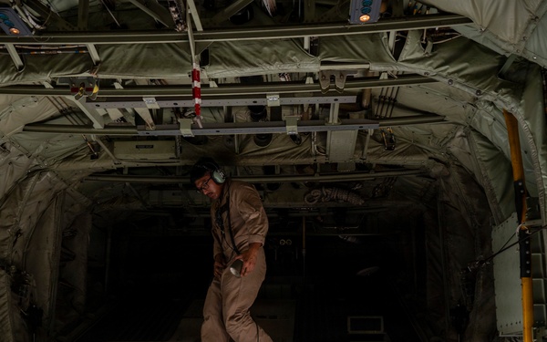 A military loadmaster or crew member in tan/khaki flight suit is inspecting the interior of a large military cargo aircraft, likely a C-130 Hercules, from the open rear ramp. The individual is wearing aviation headset and holding a flashlight while examining the empty cargo bay. The image is taken from outside the aircraft looking into the open aft cargo ramp.