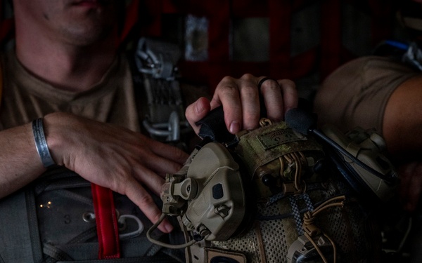 A military personnel member is handling tactical gear, specifically a helmet equipped with communications headset and multicam accessories. A patch labeled 'CRO' is clearly visible on the helmet, indicating a Combat Rescue Officer designation. The setting appears to be inside a military aircraft, with personnel wearing parachute harnesses.