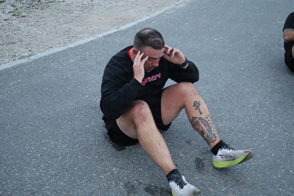 A man is sitting on an asphalt surface with his hands pressed to his temples, appearing exhausted or in distress. He is wearing a black sweatshirt with red lettering, black shorts, black socks, and white/green running shoes. He has visible tattoos on his right leg/shin area.