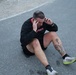 A man is sitting on an asphalt surface with his hands pressed to his temples, appearing exhausted or in distress. He is wearing a black sweatshirt with red lettering, black shorts, black socks, and white/green running shoes. He has visible tattoos on his right leg/shin area.