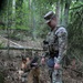 A U.S. Army Military Police (MP) soldier in MultiCam/OCP camouflage uniform is working with a military working dog, likely a Belgian Malinois or German Shepherd, in a forested environment. The soldier is wearing a tactical vest with an 'MP' patch and a unit shoulder sleeve insignia, and is holding the dog on a leash. The scene appears to be a training exercise or patrol in a woodland setting.