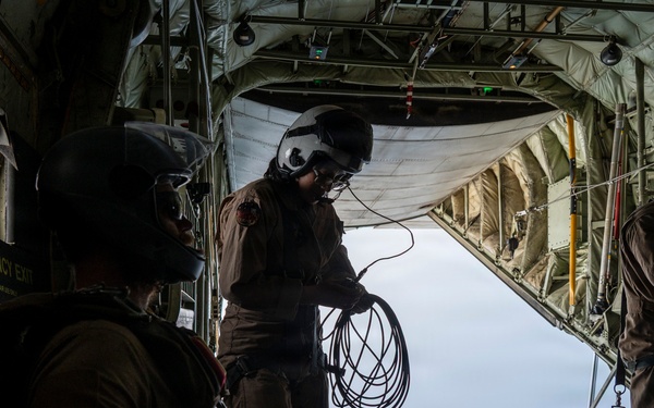 Three military personnel are positioned inside the open rear cargo ramp of a military transport aircraft, likely a C-130 Hercules, in flight. Two crew members near the open ramp are handling coiled cables or ropes, while a third individual sits toward the front of the cargo bay. The scene suggests preparation for an aerial operation such as a cargo drop or parachute deployment.