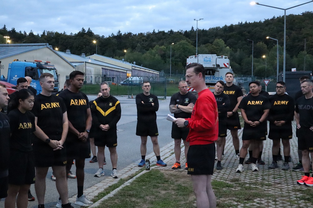 A person in a red sweatshirt appears to be addressing a group of U.S. Army soldiers wearing standard black and gold Army physical training (PT) uniforms. The gathering appears to take place outdoors on a military installation during early morning or dusk hours, with street lights illuminated. The setting appears to be a European military base, suggested by the road signage and architectural style visible in the background.