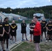 A person in a red sweatshirt appears to be addressing a group of U.S. Army soldiers wearing standard black and gold Army physical training (PT) uniforms. The gathering appears to take place outdoors on a military installation during early morning or dusk hours, with street lights illuminated. The setting appears to be a European military base, suggested by the road signage and architectural style visible in the background.