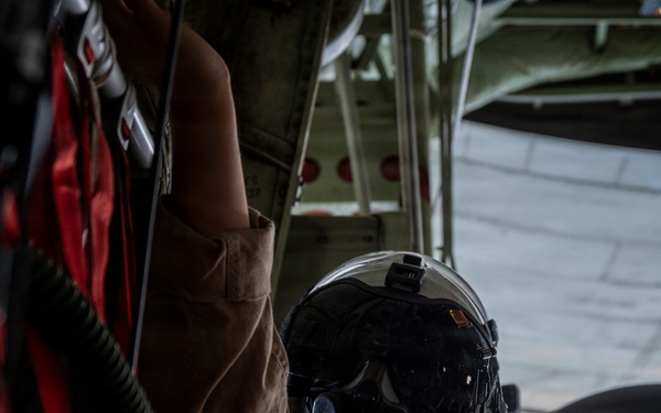A military aircrew member wearing a flight helmet with oxygen mask and visor is seated inside a military aircraft, likely a transport or tanker aircraft. The individual is reaching upward toward overhead equipment or controls, wearing a tan/brown flight suit. A second helmeted crew member is partially visible in the foreground.