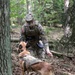 A U.S. Army Military Police (MP) soldier in MultiCam/OCP camouflage uniform interacts with a military working dog (MWD) in a wooded forest setting. The dog, wearing a tactical camouflage vest with a name tag reading 'LUCKY', appears to be a Belgian Malinois. The scene likely depicts a K9 handler training exercise or operational patrol.