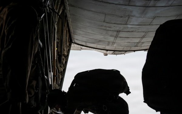 The image shows silhouetted military personnel standing at the open rear ramp of a military cargo aircraft, likely a C-130 or similar transport. One individual is leaning forward and looking out of the open ramp toward the sky, suggesting a pre-jump inspection or aerial observation. The strong backlight from the open ramp creates a high-contrast silhouette effect.