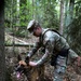 A U.S. Army Military Police (MP) soldier in OCP (Operational Camouflage Pattern) uniform interacts with a military working dog, likely a Belgian Malinois, in a forested environment. The soldier is bending down to handle or reward the dog, which appears alert with its tongue out. This appears to be a K9 unit training or operational exercise in a woodland setting.