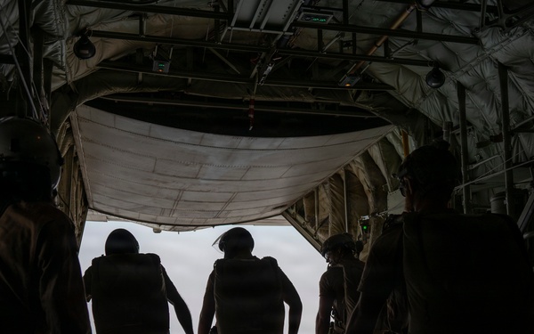 Several military personnel in silhouette are standing at the open rear ramp of a large military cargo aircraft, likely a C-130 Hercules or similar transport, in flight. The figures are wearing tactical gear including helmets, body armor/parachute packs, and combat uniforms. The scene appears to depict a pre-jump or airdrop operation with overcast skies visible through the open ramp.