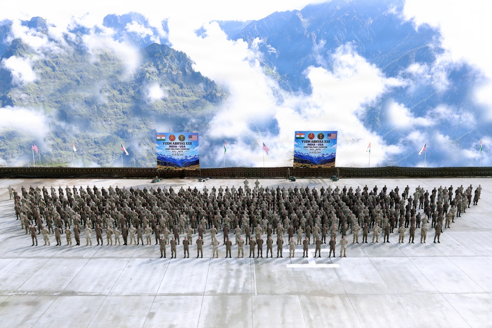 This is a large group photograph taken during 'Yudh Abhyas XXII', a joint military exercise between India and the USA. Hundreds of military personnel from both nations are assembled in formation on a parade ground, with two identical event banners prominently displayed in the background. The setting appears to be a mountainous region, consistent with high-altitude military training locations in India.