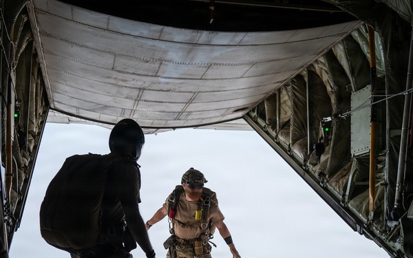 Military personnel are conducting a parachute jump operation from the open rear ramp of a large military cargo aircraft, likely a C-17 or C-130. One jumper is already airborne just outside the aircraft, while a second personnel member approaches the ramp edge preparing to jump. At least two additional personnel are visible inside the aircraft near the sides of the ramp.