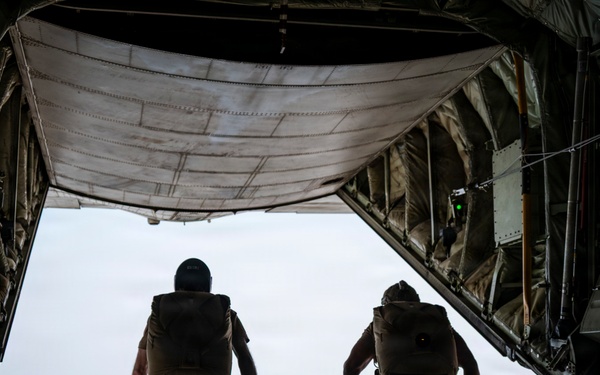 Two military personnel in desert/tan camouflage uniforms and parachute packs are standing at the open rear ramp of a military cargo aircraft in flight. They appear to be preparing for a parachute jump, positioned at the edge of the open ramp with sky visible beyond. The image is taken from inside the aircraft looking toward the open ramp door.