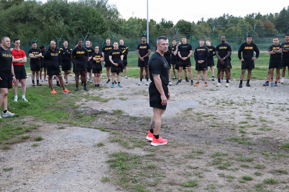 A group of U.S. Army soldiers in physical training (PT) uniforms are assembled outdoors in a semicircle formation. A central figure, appearing to be a senior non-commissioned officer or instructor, stands facing the group in a leadership position. The setting appears to be a military installation or training area with a chain-link fence and trees in the background.