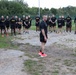 A group of U.S. Army soldiers in physical training (PT) uniforms are assembled outdoors in a semicircle formation. A central figure, appearing to be a senior non-commissioned officer or instructor, stands facing the group in a leadership position. The setting appears to be a military installation or training area with a chain-link fence and trees in the background.