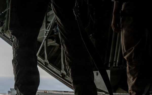 The image shows the interior of a military cargo aircraft, likely a C-130 Hercules, with the rear ramp open revealing sky and water below. Two or more personnel in military uniforms are standing on the cargo ramp floor, appearing to handle straps or rigging equipment. The scene suggests preparation for an airdrop, cargo deployment, or parachute operation.